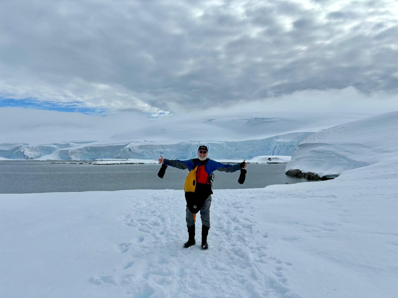 Drew standing on snow in Antarctica with arms outstretched and thumbs up, glaciers in the background