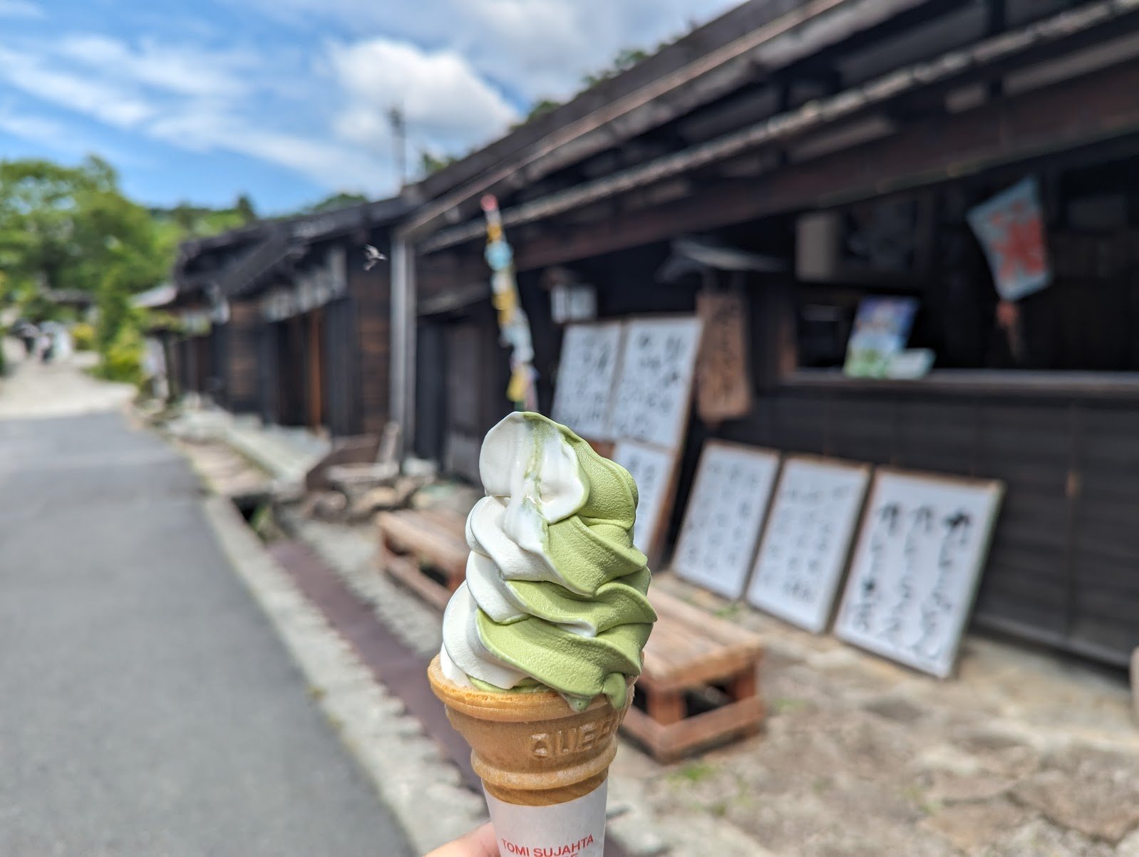 A matcha and vanilla swirl soft-serve cone held up in front of the dark timber post-town buildings on the Nakasendo trail — June 2024
