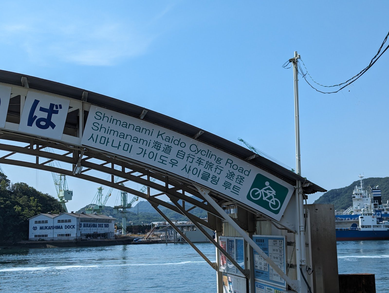 The Shimanami Kaido Cycling Road archway at the Onomichi ferry crossing, with the Mukaishima shipyard behind — June 2024