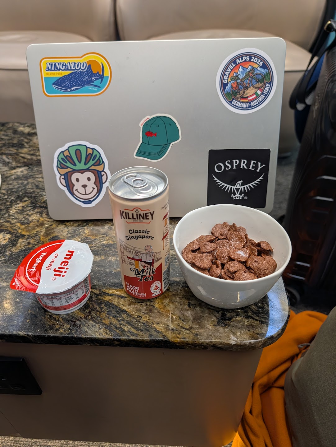 Bowl of chocolate cereal, Meiji yoghurt and Killiney milk tea on a marble table next to a sticker-covered laptop at SATS Premier Lounge