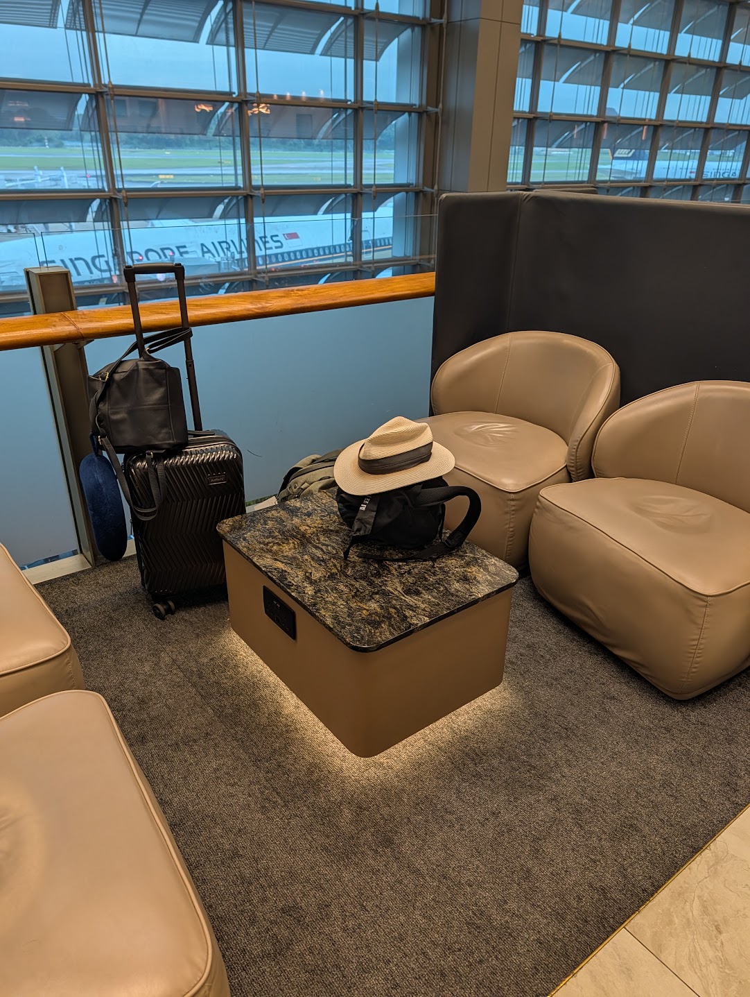 Lounge armchairs and a marble side table by a window overlooking a Singapore Airlines aircraft on the tarmac at Changi Terminal 3