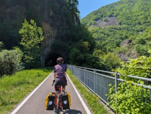 Cyclist on EuroVelo 7 approaching an alpine tunnel, fully loaded panniers, northern Italy, April 2026