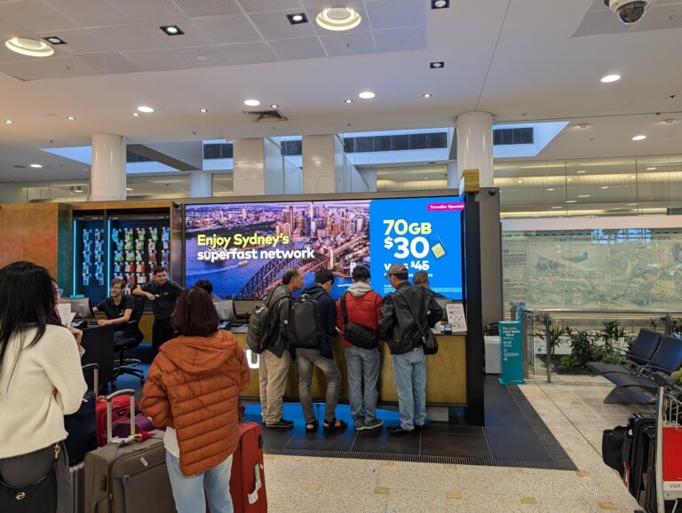 Travellers queue at the Optus SIM counter at Sydney Airport — why an Airalo travel eSIM beats airport roaming. Points Brotherhood.