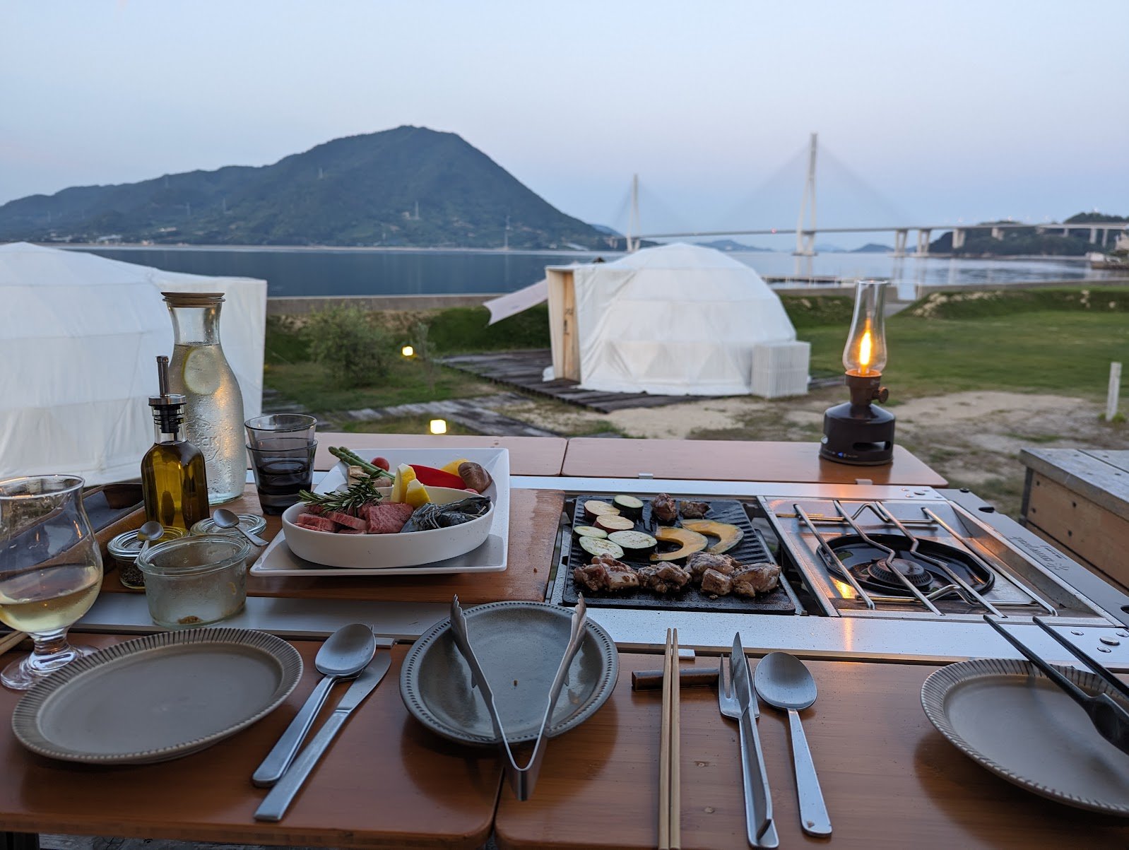 A BBQ dinner set up at dusk at WAKKA, with the Seto Inland Sea, a bridge and dome tents in the background — June 2024