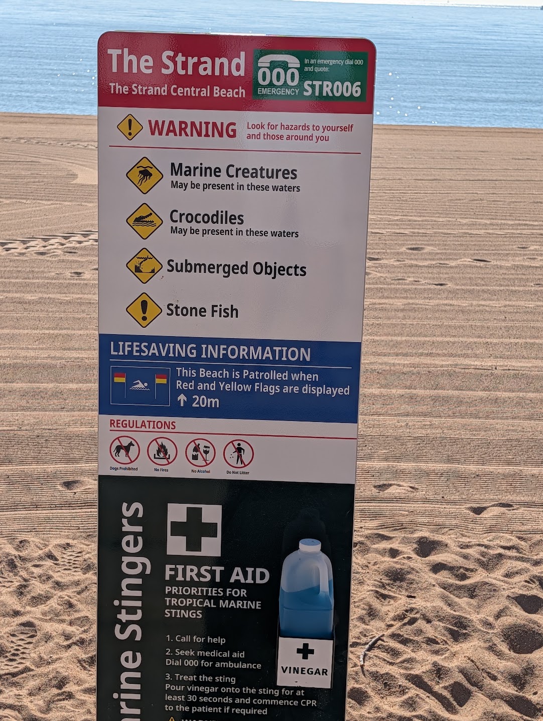 Beach safety warning sign at The Strand in Townsville listing marine stingers, crocodiles, submerged objects and stonefish hazards